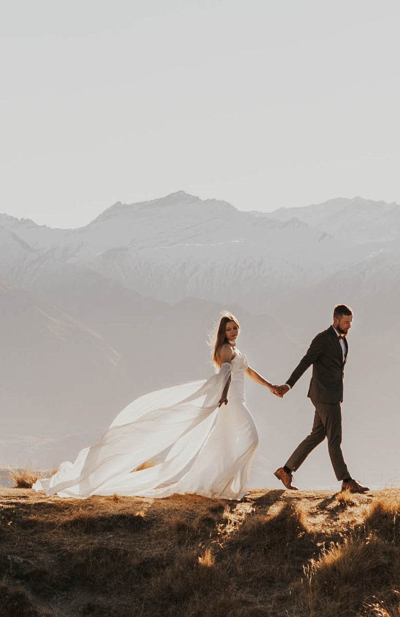 Couple holding hands on a hilltop, chiffon sleeves and train of a silk wedding dress sweeping the ground, FEYRE