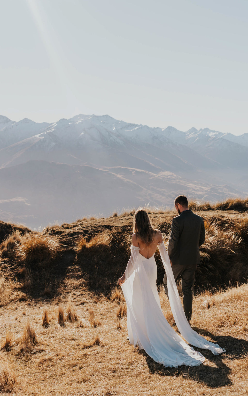Back view of a couple walking into the hills, open back silk wedding dress and chiffon sleeves trailing, FEYRE