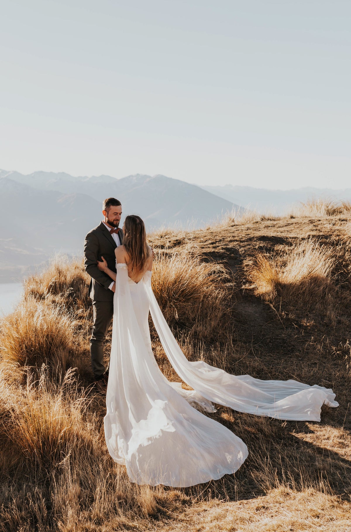 Bride embracing the groom on golden tussock, silk wedding dress with long chiffon sleeves across the grass, FEYRE