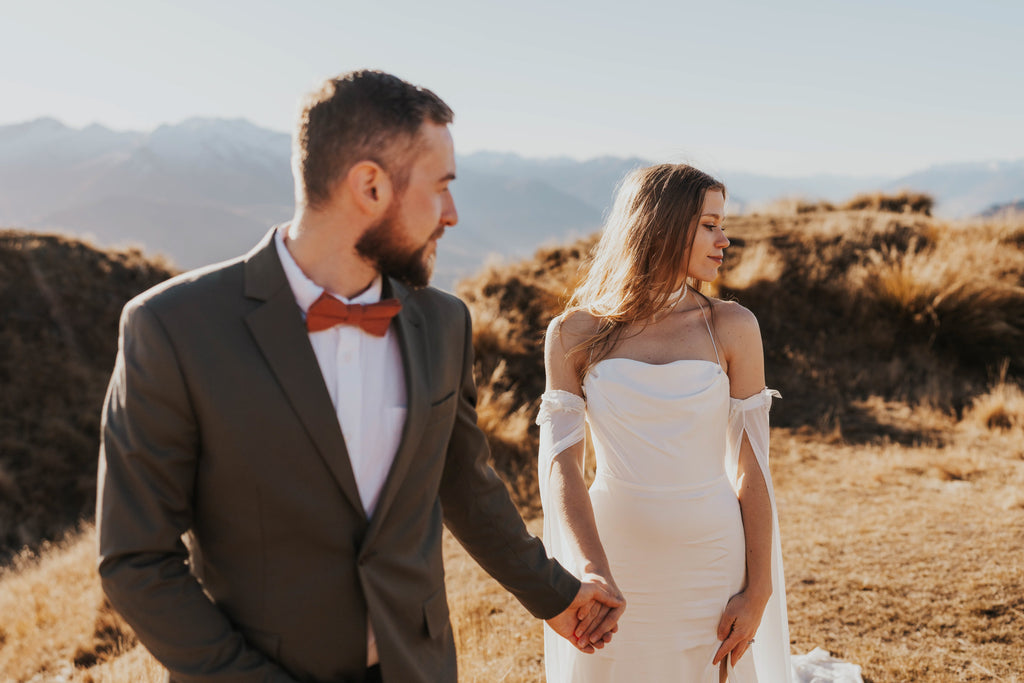 Couple holding hands in golden light, bride in a fitted silk wedding dress with detachable sleeves at the arm, FEYRE
