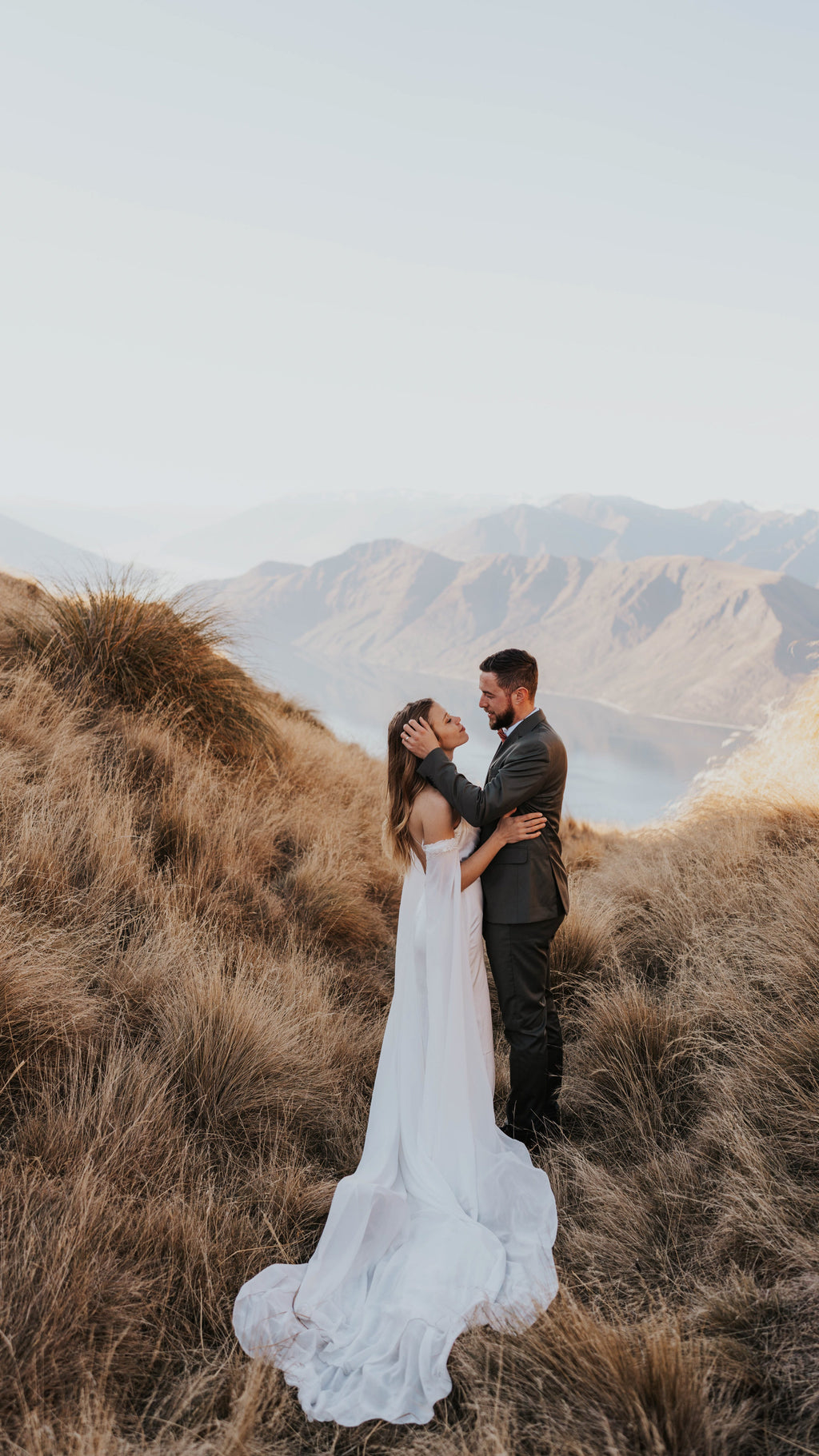 Bride and groom embracing on a grassy slope above a lake, silk wedding dress pooling into a soft train, FEYRE