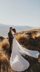 Couple touching foreheads on a mountain ridge, bride in a silk wedding dress with chiffon sleeves blowing out behind, FEYRE
