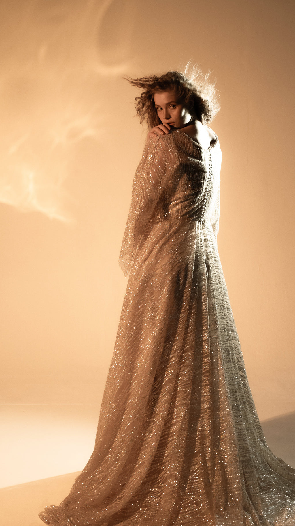 Model glancing over her shoulder, sheer lace sleeve gathered at her chin, long champagne wedding dress trailing behind, Galaxy Champagne 2.0