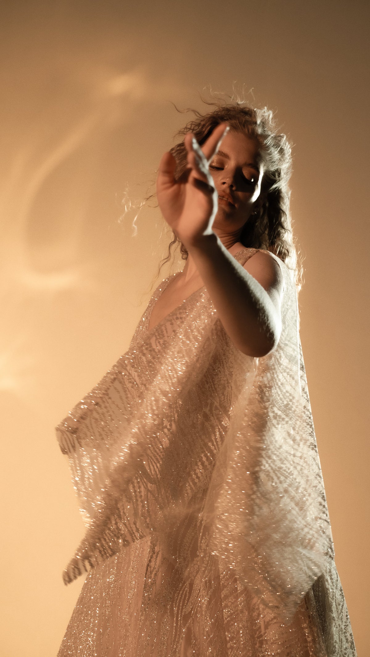 Model reaching toward the camera, slit sleeve of a glittering champagne lace wedding dress floating around the arm, Galaxy Champagne 2.0
