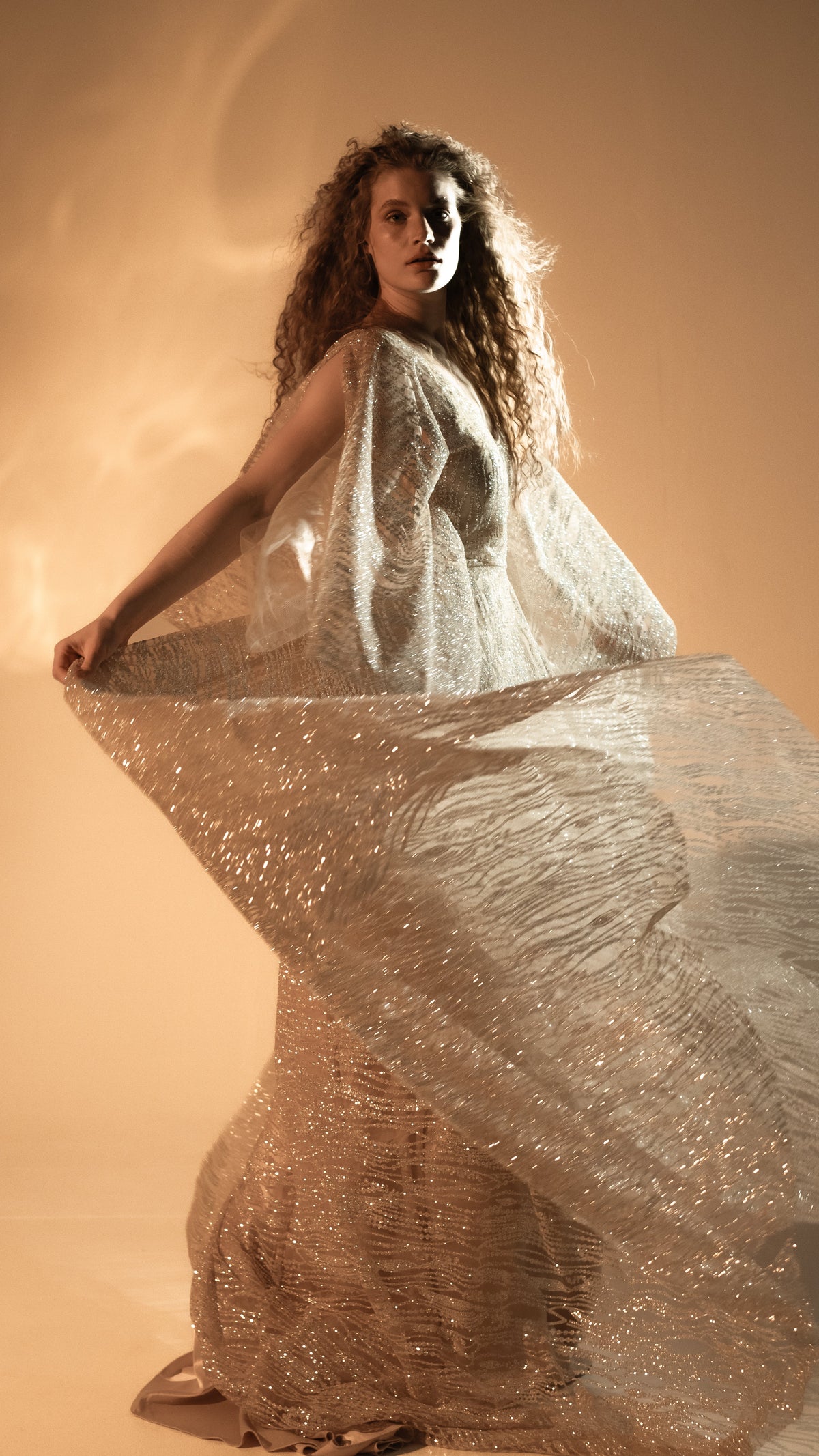 Model turning with a wide sheer sleeve lifted, champagne glitter lace wedding dress catching the amber light, Galaxy Champagne 2.0