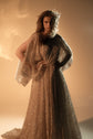 Hand on hip in a long champagne glitter lace wedding dress, cape-like sleeve falling open at the arm, Galaxy Champagne 2.0