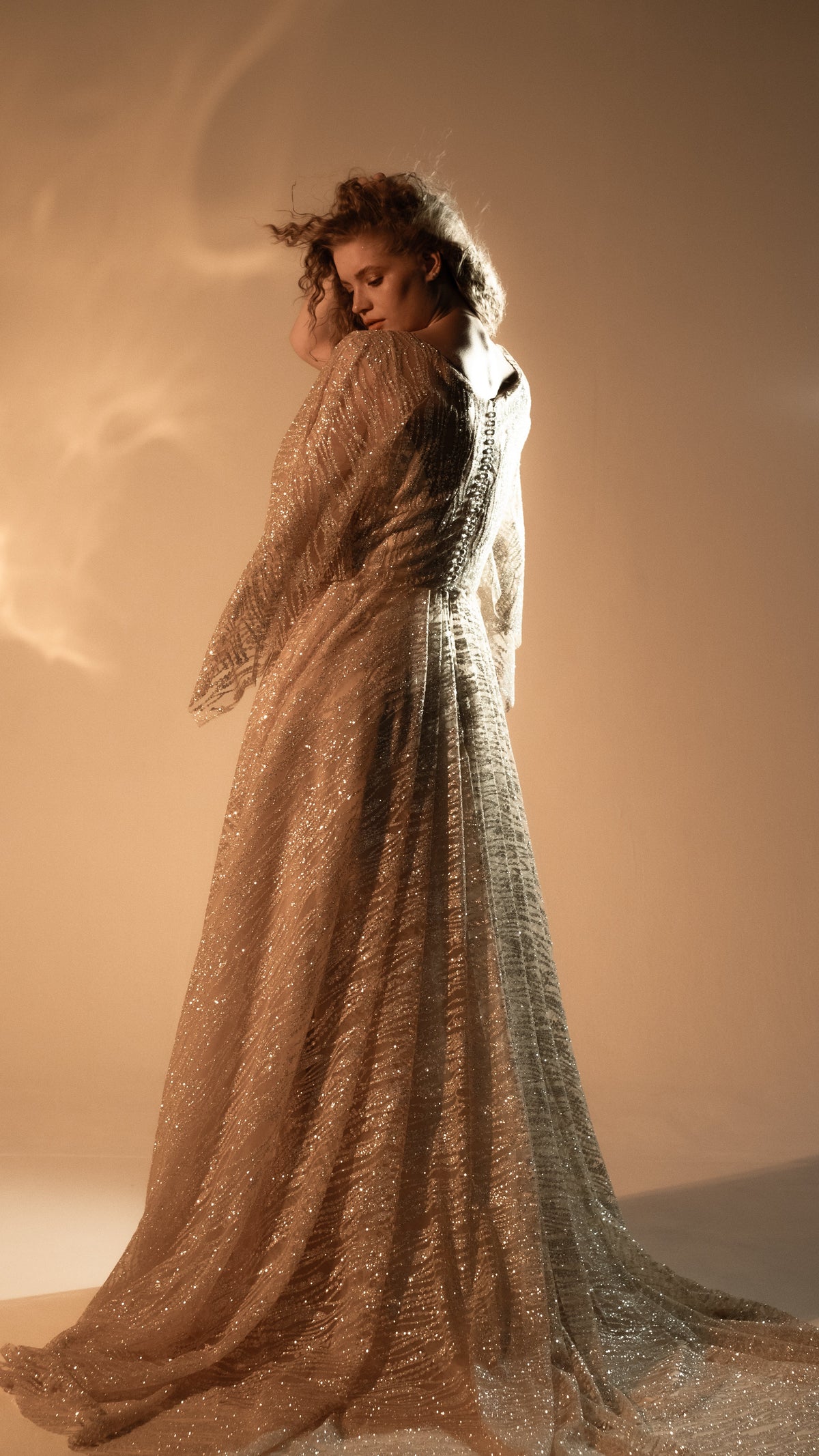 Three-quarter back view of a champagne lace wedding dress with a sweeping train and button back, model looking down, Galaxy Champagne 2.0