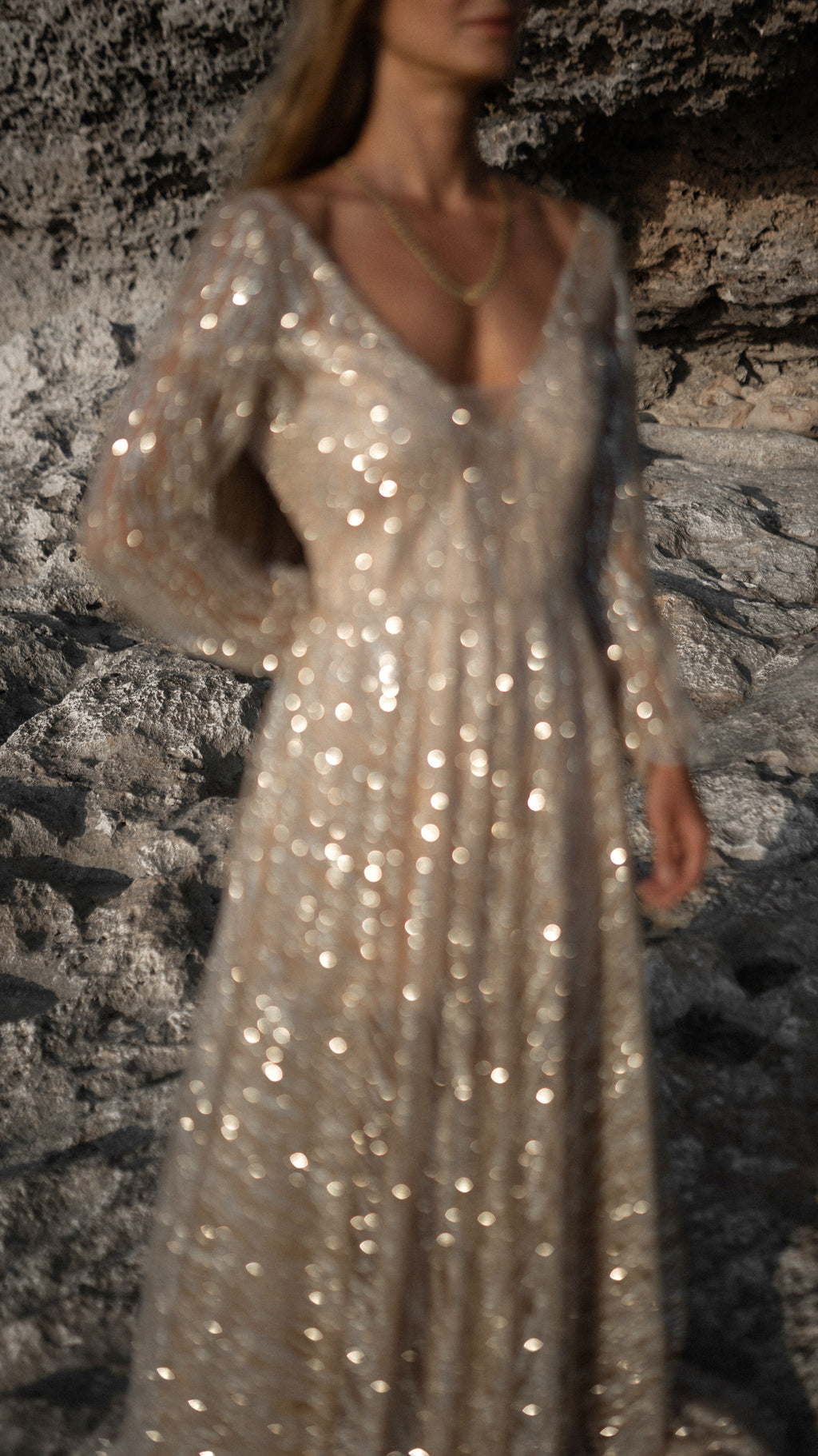 Champagne glitter lace wedding dress with a deep V neckline and sheer long sleeves, sparkling against a dark rock wall. Galaxy Champagne by Ephemeris