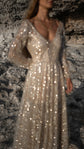 Champagne glitter lace wedding dress with a deep V neckline and sheer long sleeves, sparkling against a dark rock wall. Galaxy Champagne by Ephemeris