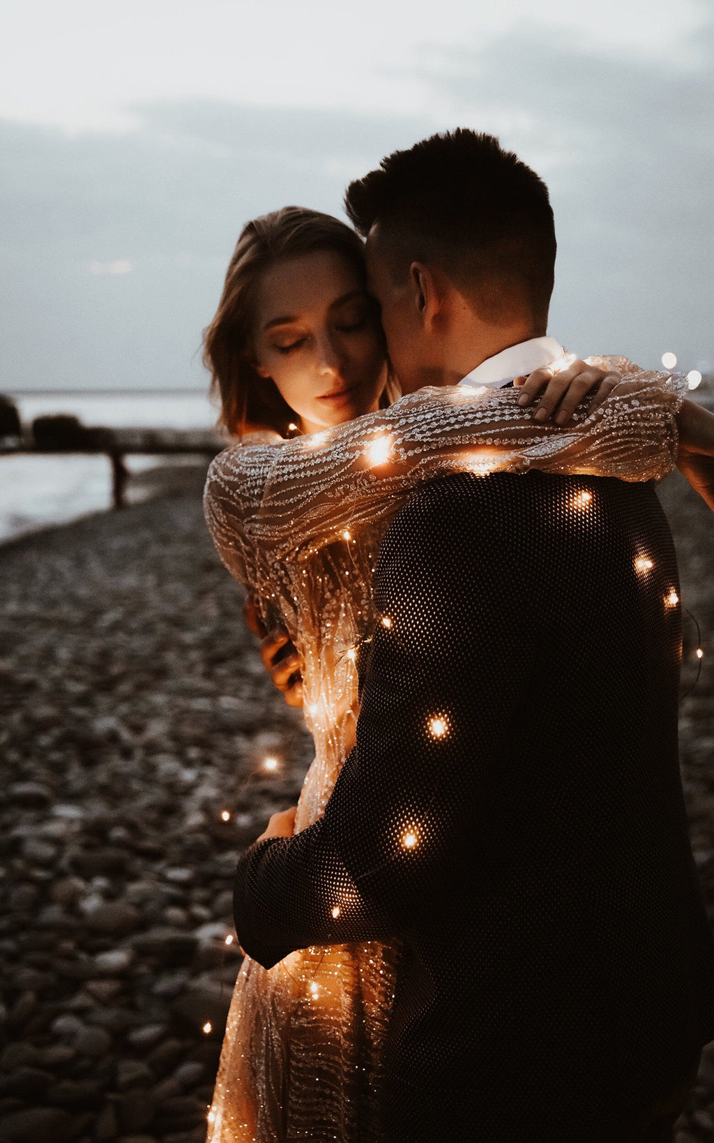 Close embrace at dusk on the shore, sheer glitter lace sleeve of a champagne wedding dress lit by tiny lights, Galaxy Champagne