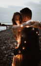 Close embrace at dusk on the shore, sheer glitter lace sleeve of a champagne wedding dress lit by tiny lights, Galaxy Champagne