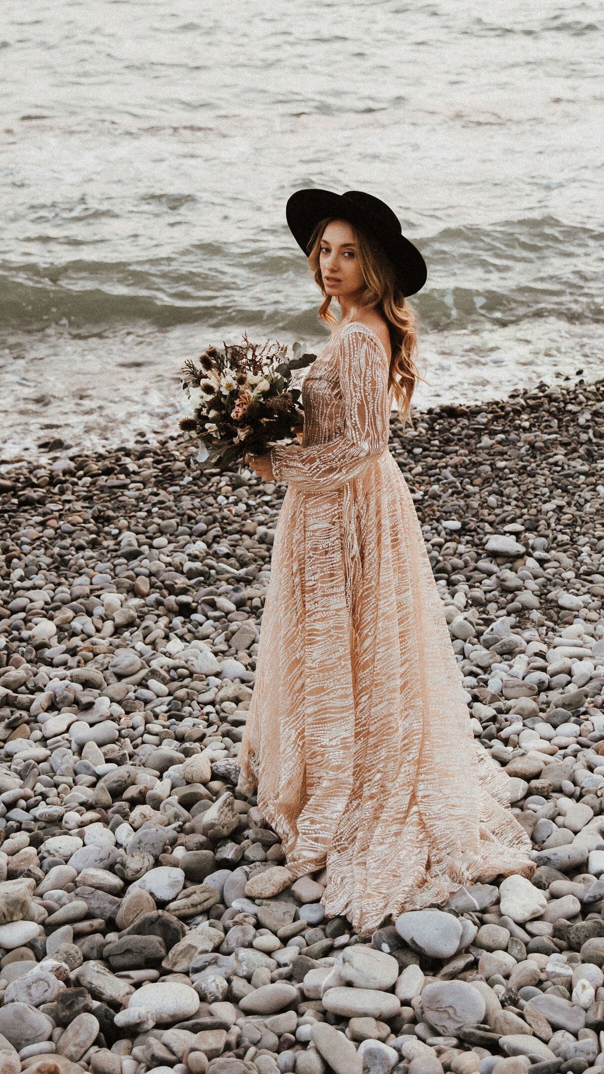 Boho bride on a pebble beach holding a dried flower bouquet, champagne glitter lace wedding dress and black hat, Galaxy Champagne