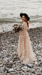 Boho bride on a pebble beach holding a dried flower bouquet, champagne glitter lace wedding dress and black hat, Galaxy Champagne