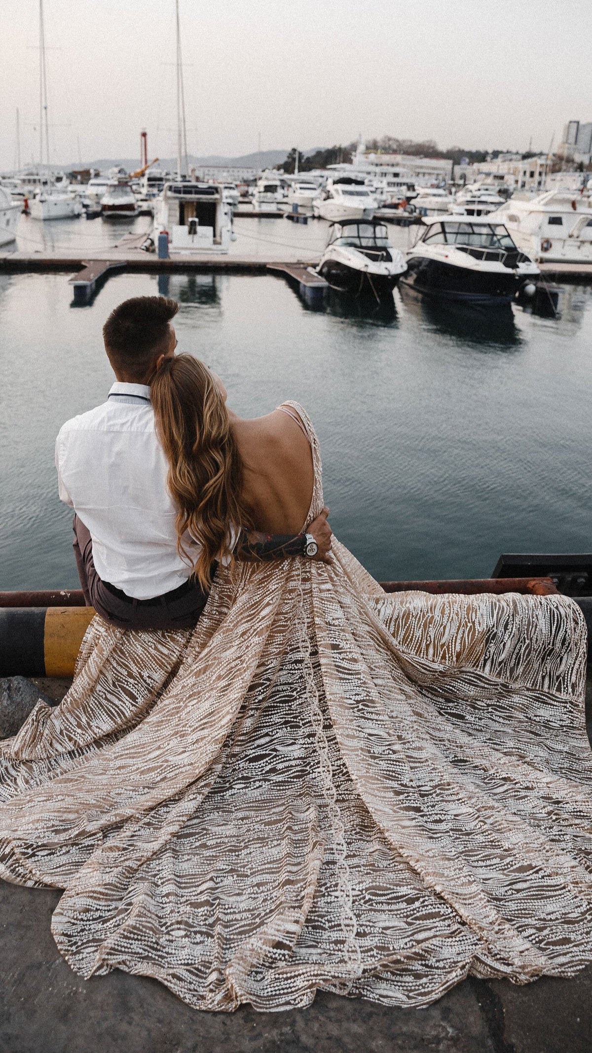 Back view of a backless champagne wedding dress with a wide train spread over a marina pier, couple seated by the boats, Galaxy Champagne