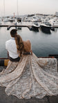 Back view of a backless champagne wedding dress with a wide train spread over a marina pier, couple seated by the boats, Galaxy Champagne