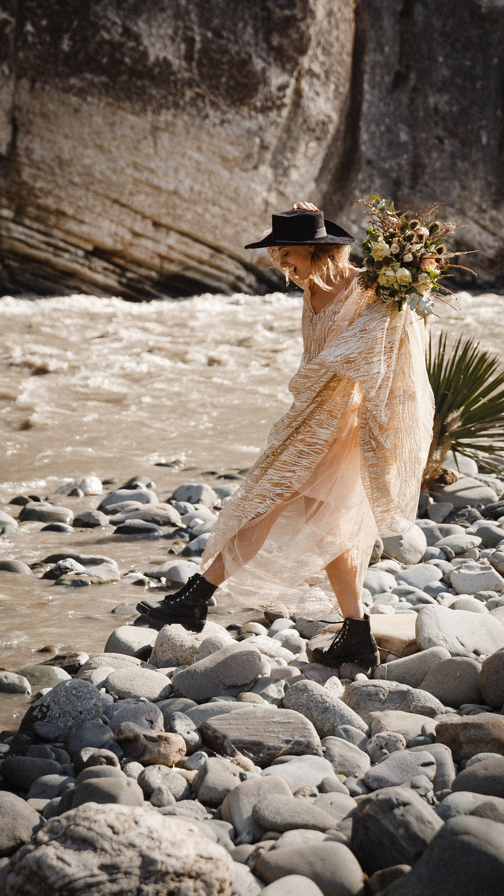 Bride in boots stepping across river stones in a champagne lace wedding dress, holding a hat and wild bouquet, cliffs behind, Galaxy Champagne