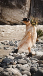 Bride in boots stepping across river stones in a champagne lace wedding dress, holding a hat and wild bouquet, cliffs behind, Galaxy Champagne