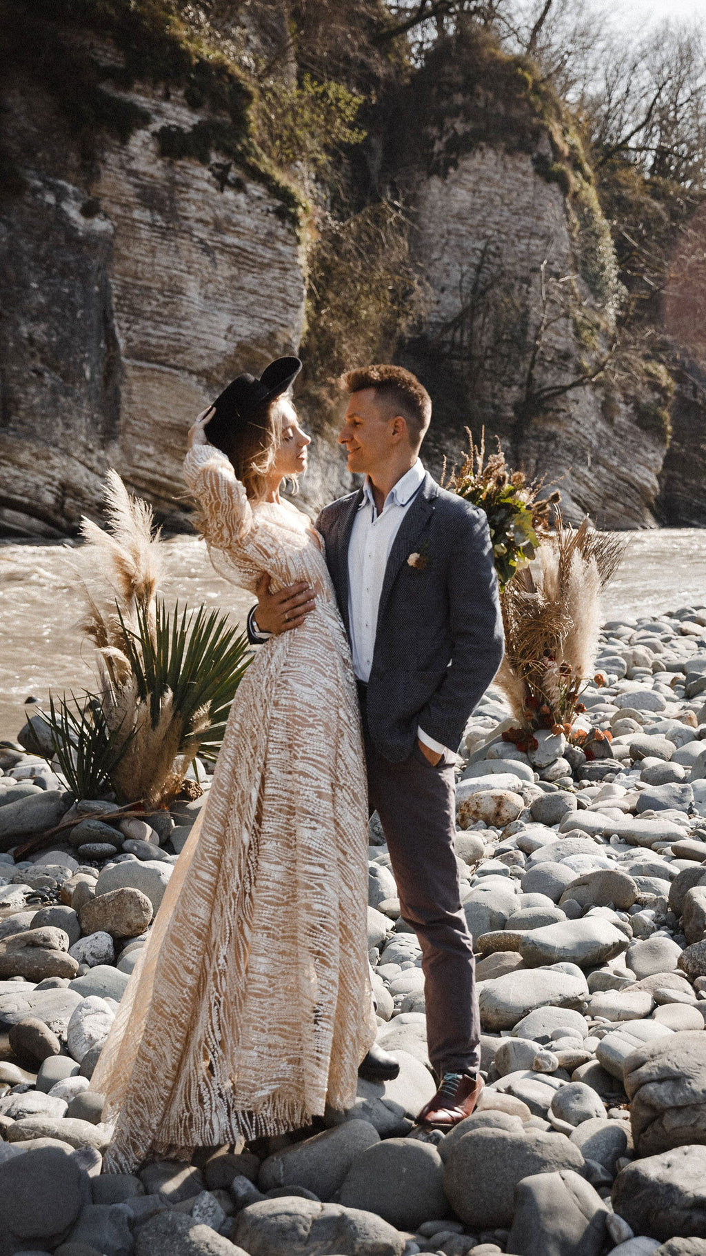Elopement couple among river stones and pampas grass, bride in a long champagne glitter lace wedding dress and hat, Galaxy Champagne