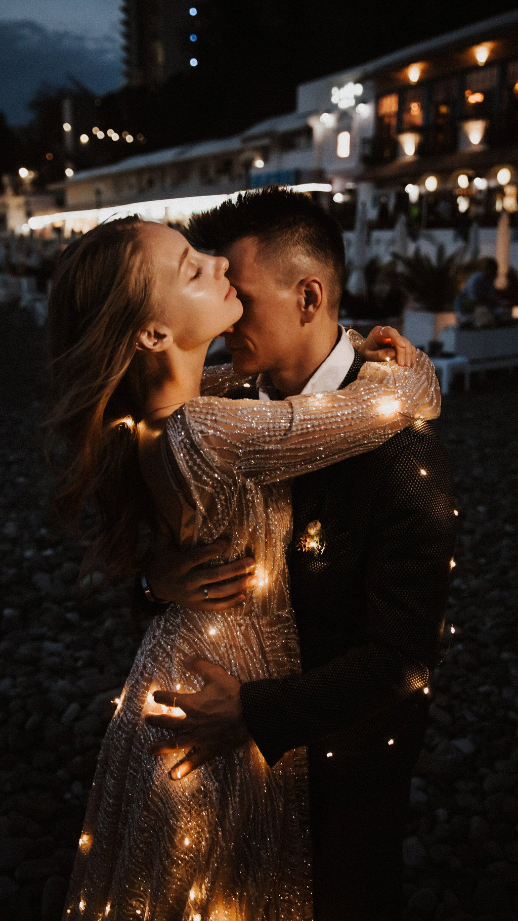 Kiss under fairy lights at night, sheer long sleeves of a champagne glitter wedding dress wrapped around the groom, Galaxy Champagne