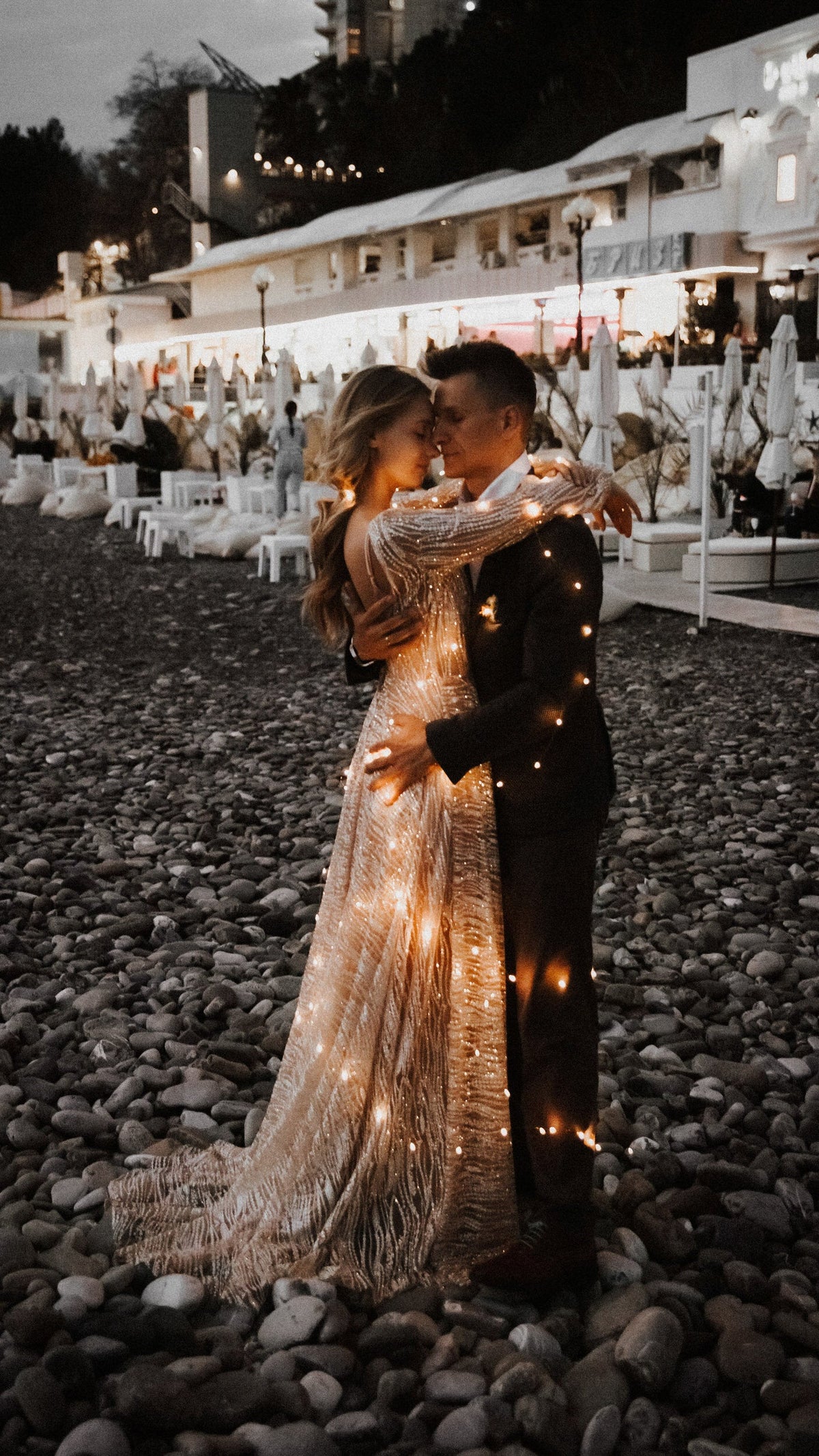 Couple hugging on a dark pebble beach, lights glinting through the skirt and train of a champagne lace wedding dress, Galaxy Champagne