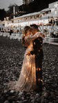 Couple hugging on a dark pebble beach, lights glinting through the skirt and train of a champagne lace wedding dress, Galaxy Champagne