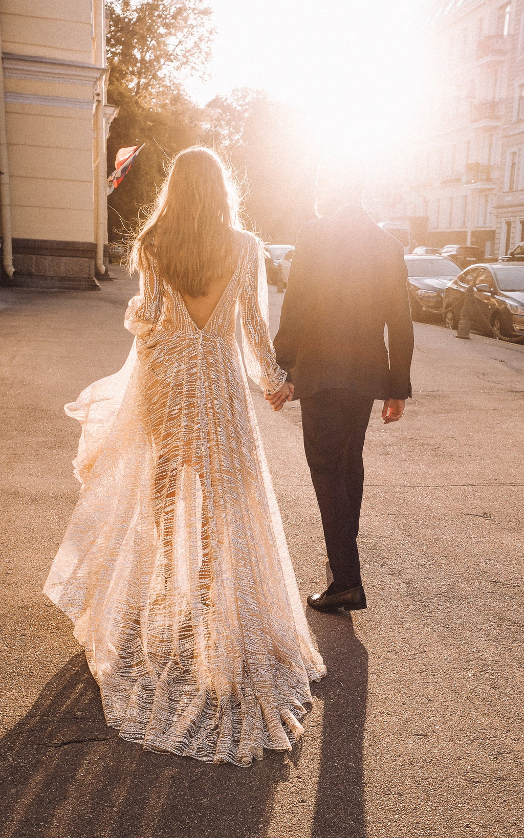 Back view of a low V-back champagne lace wedding dress backlit by the sun as the couple walks down a city street, Galaxy Champagne