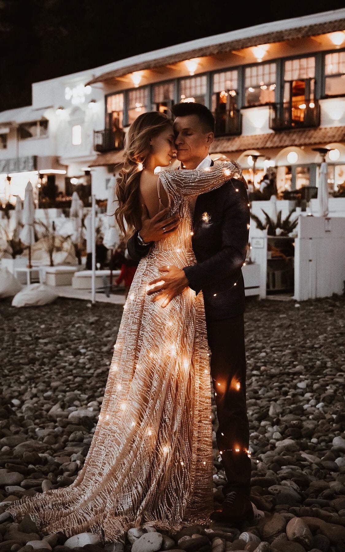 Open-back champagne lace wedding dress wrapped in fairy lights as the couple embraces on a pebble beach at night, Galaxy Champagne