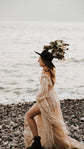 Side view of a sparkly beige wedding dress with a front slit on a pebble shore by the sea, bride in boots and a flower-trimmed hat, Galaxy Champagne