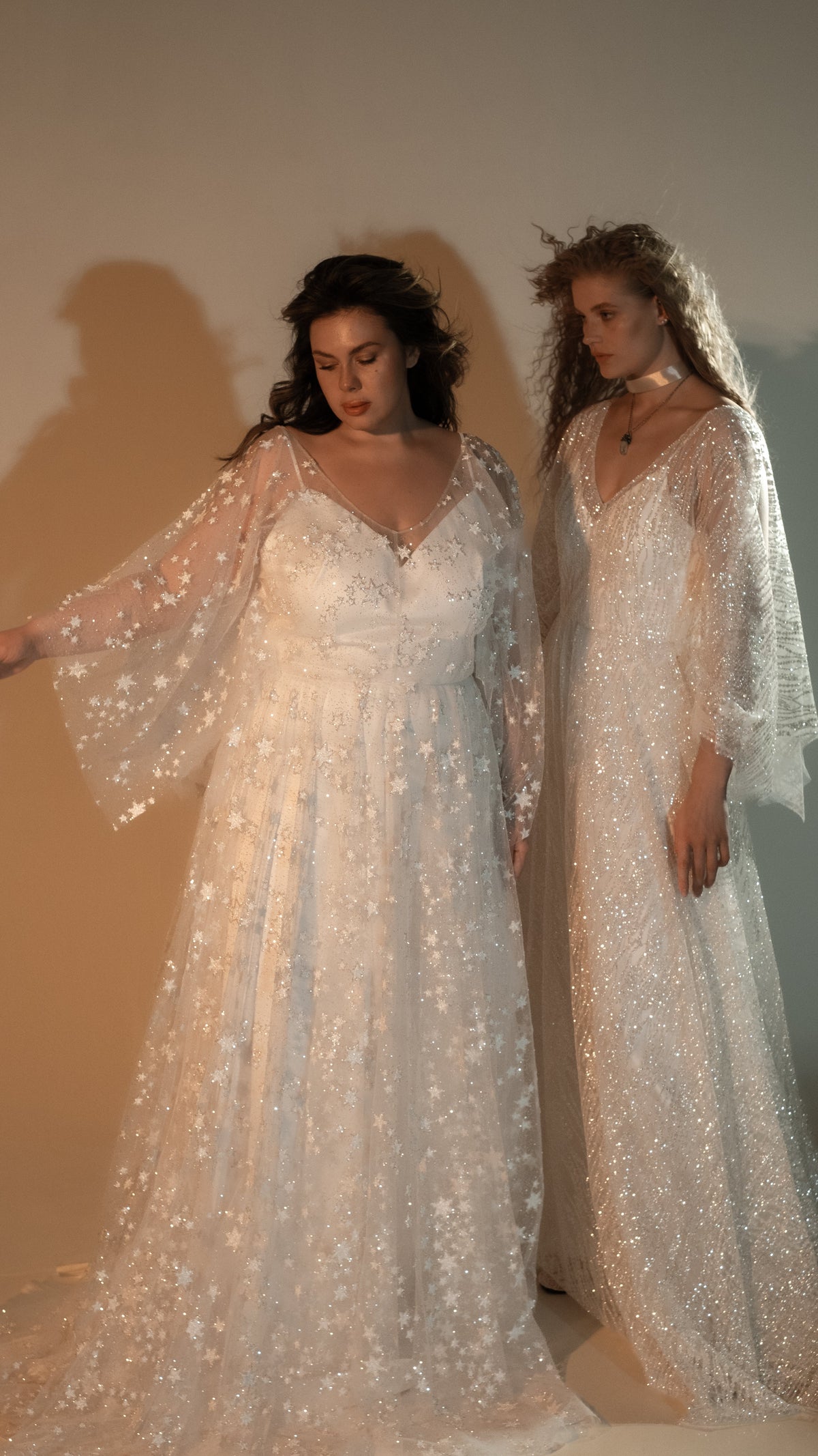 Two models side by side, a star lace wedding dress with flutter sleeves and an ivory glitter lace wedding dress with a neck scarf, Galaxy Ivory 2.0