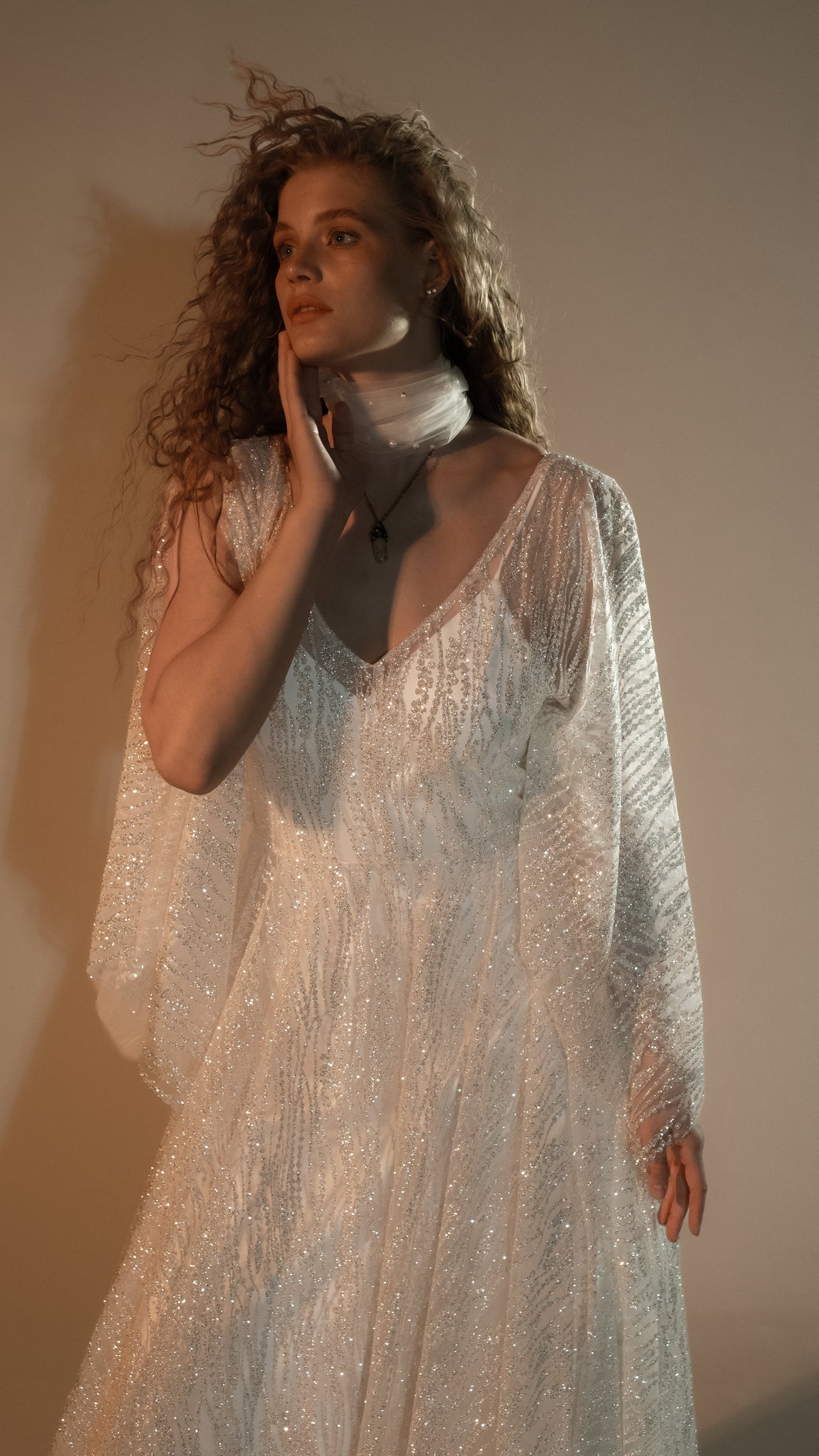Waist-up crop of an ivory sparkle lace wedding dress with sheer cape-like sleeves, tulle wrapped at the neck, hand at the cheek, Galaxy Ivory 2.0