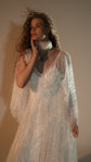 Waist-up crop of an ivory sparkle lace wedding dress with sheer cape-like sleeves, tulle wrapped at the neck, hand at the cheek, Galaxy Ivory 2.0