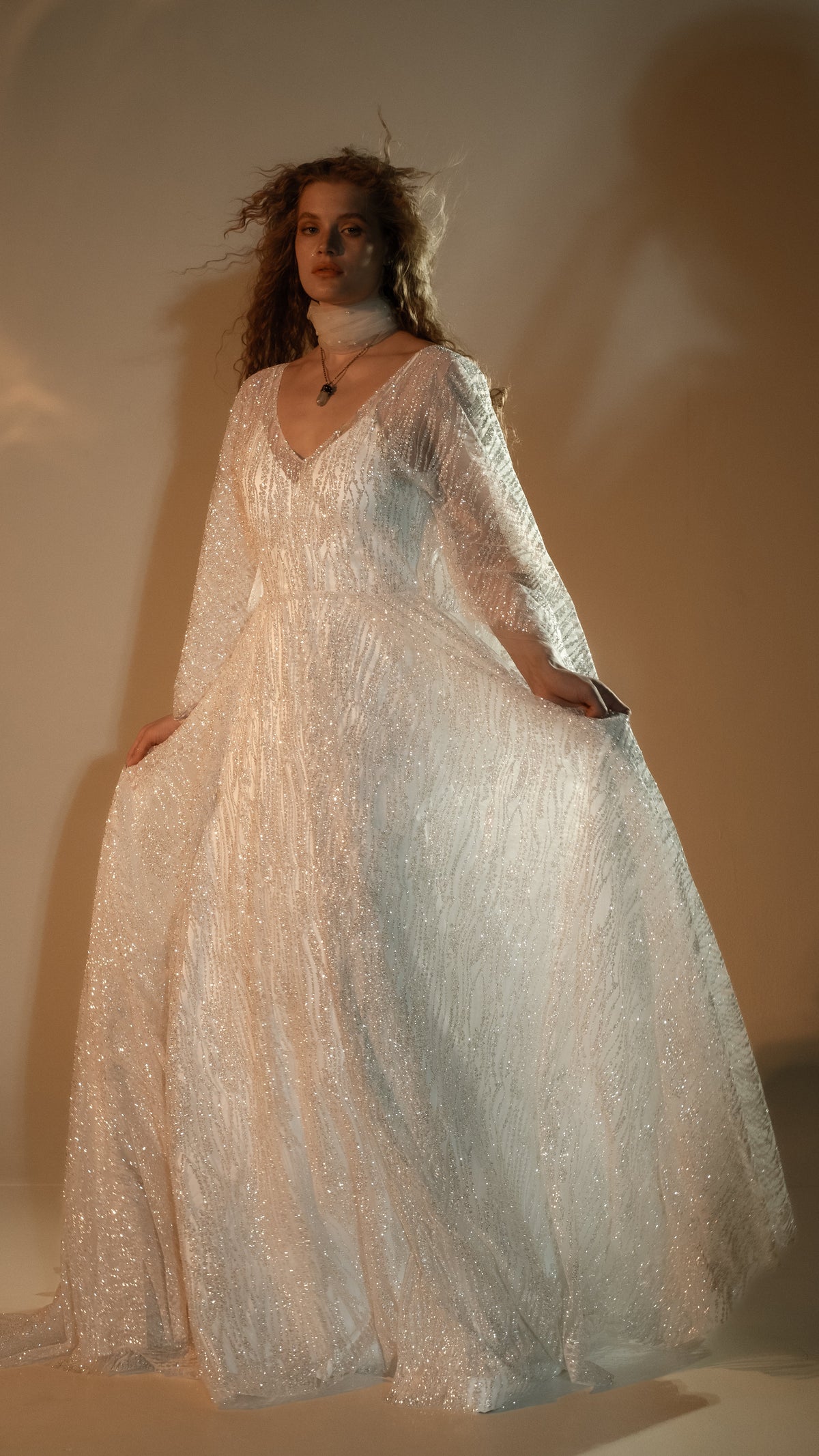 Model holding out the full skirt of a V-neck ivory glitter lace wedding dress, sleeves falling open at the arms, Galaxy Ivory 2.0