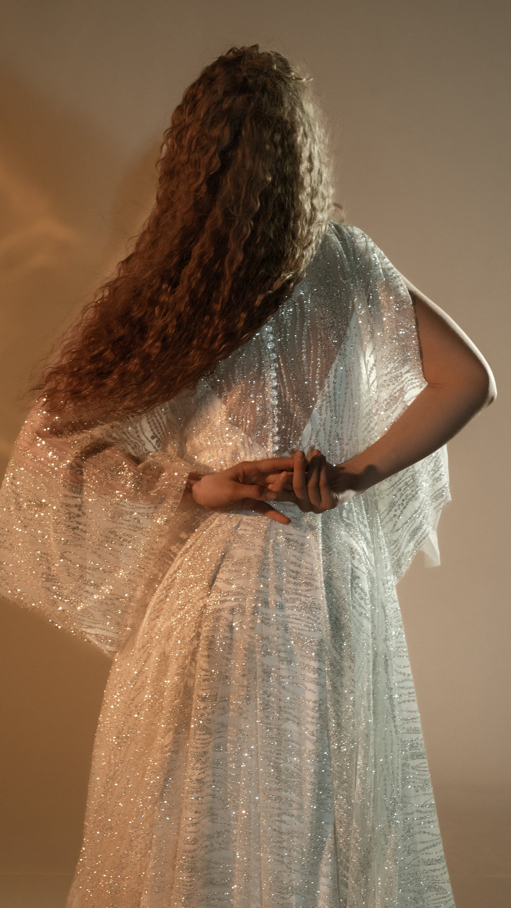 Back view of an ivory lace wedding dress with a sheer tulle back and button row, hands clasped behind, long sleeve draped, Galaxy Ivory 2.0