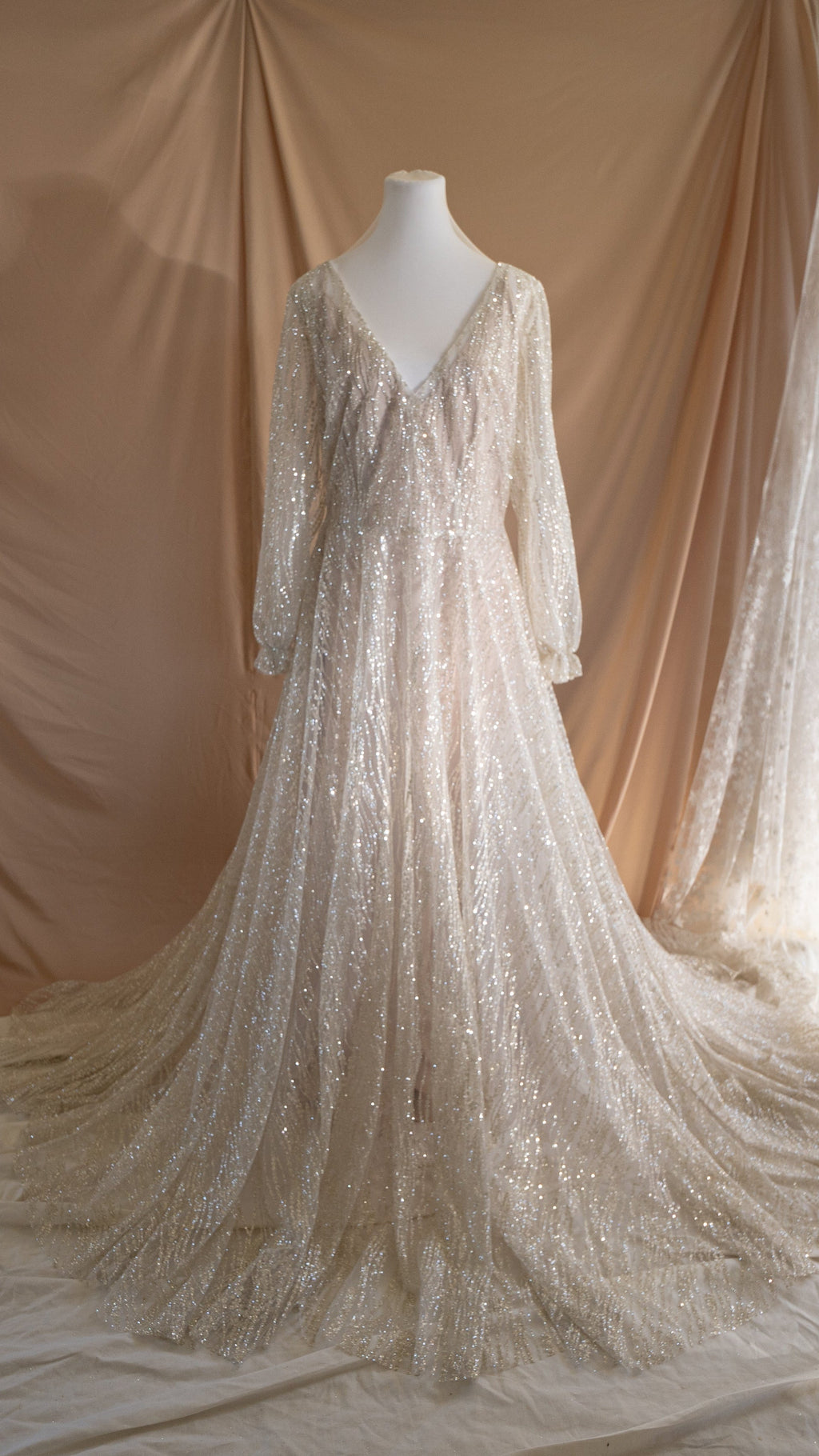Sample wedding dress in ivory glitter lace on a dress form, V neckline, long sleeves and a wide train on a beige backdrop. Galaxy Ivory Sample by Ephemeris