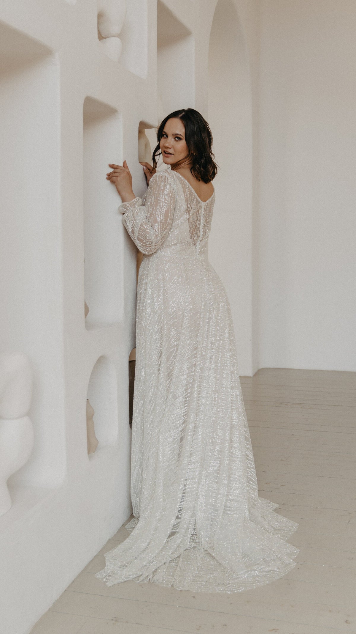 Bride against a sculpted white wall looking back, scoop back with buttons on an ivory lace wedding dress and a soft train, Galaxy Ivory Sample