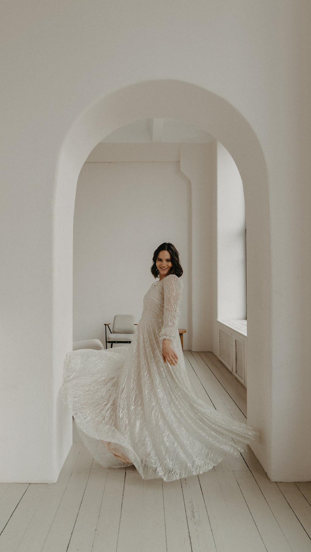 Bride mid-spin in a white archway, glancing back over her shoulder, long-sleeve ivory lace wedding dress fanning out, Galaxy Ivory Sample