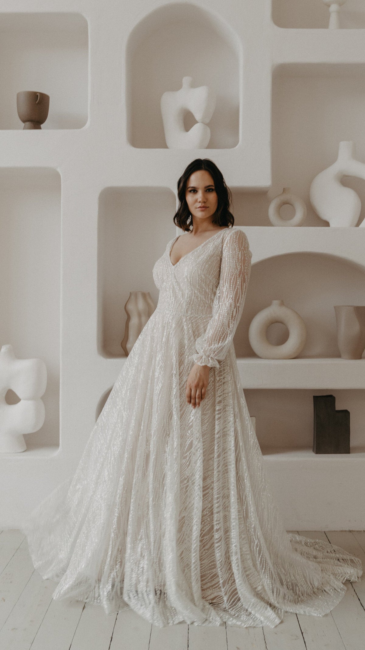 Bride before white shelves of ceramics in a V-neck ivory glitter lace wedding dress with cuffed long sleeves, Galaxy Ivory Sample