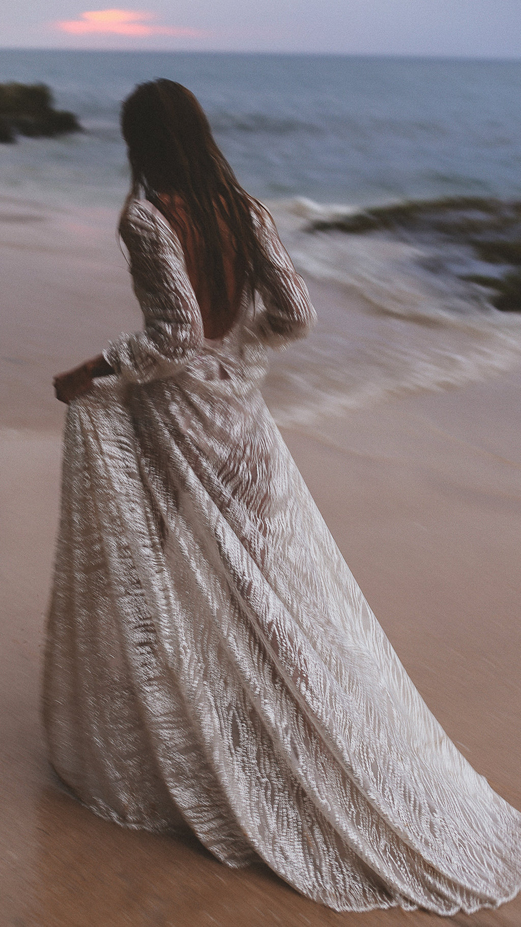 Back view of an ivory lace wedding dress with an open back on a beach at dusk, skirt lifted above the waves, Galaxy Ivory Sample