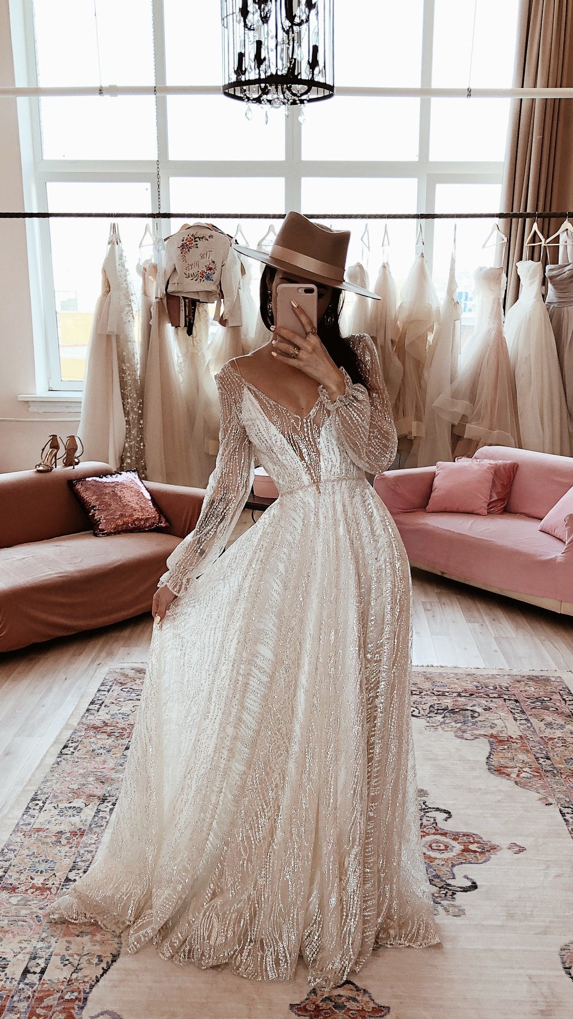 Ivory glitter lace wedding dress with a V neckline and sheer long sleeves, bride taking a mirror selfie in a bridal showroom. Galaxy Ivory by Ephemeris