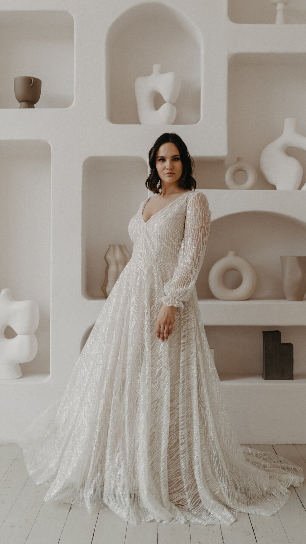 Front view of a V-neck ivory lace wedding dress with gathered cuffs before white shelves of ceramic vases, Galaxy Ivory