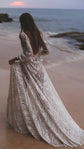 Back view of an open-back ivory lace wedding dress on a beach at dusk, bride lifting the long skirt over wet sand, Galaxy Ivory