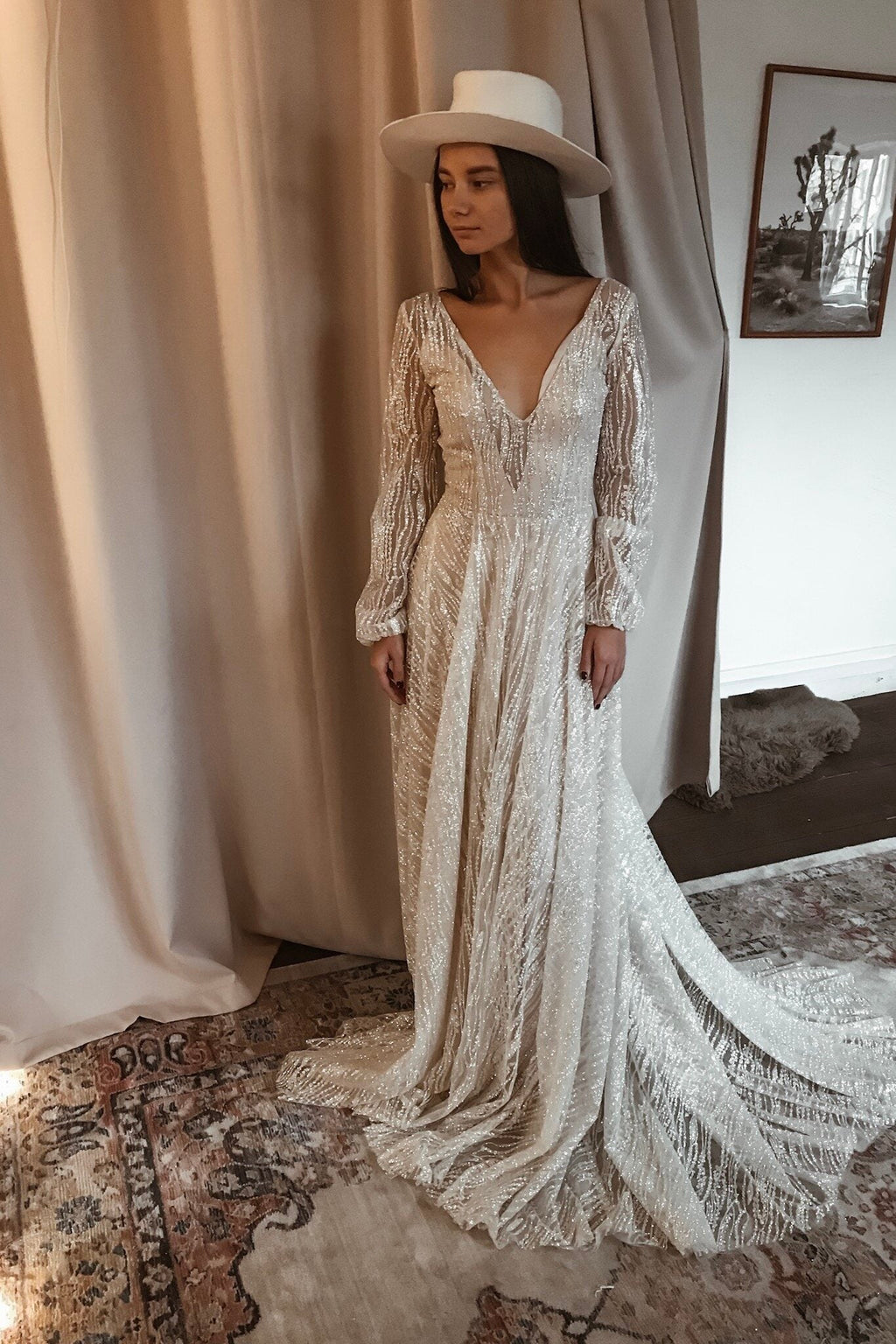 Ivory lace wedding dress with a deep V neckline and long cuffed sleeves, bride in a wide-brim hat on a patterned rug, Galaxy Ivory