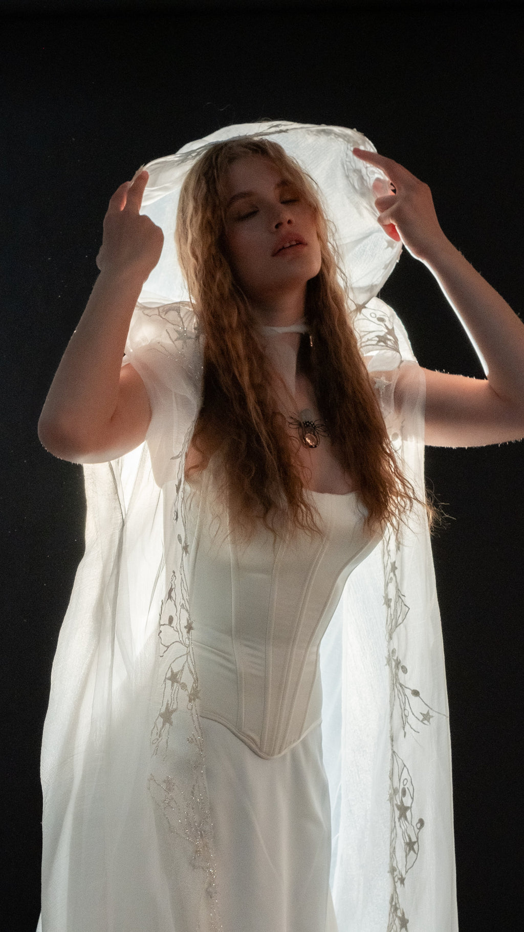 Hooded bridal cape in ivory crinkle chiffon with star vine lace edging, raised over the head above a white corset gown. Holly Cape by Ephemeris