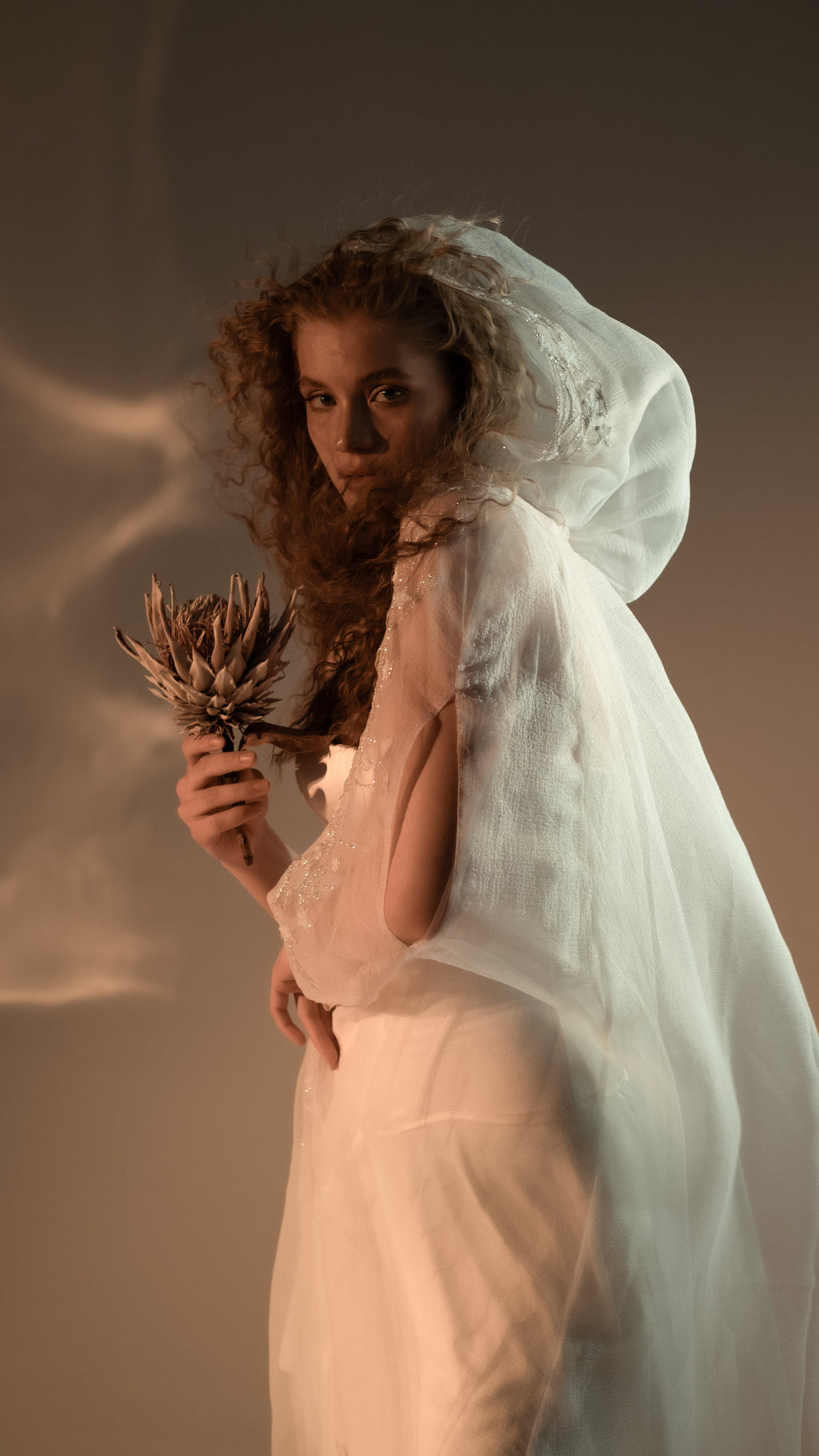 Side view of an ivory hooded bridal cape with soft crinkle chiffon folds over a long white gown, dried protea in hand, Holly Cape