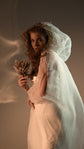 Side view of an ivory hooded bridal cape with soft crinkle chiffon folds over a long white gown, dried protea in hand, Holly Cape