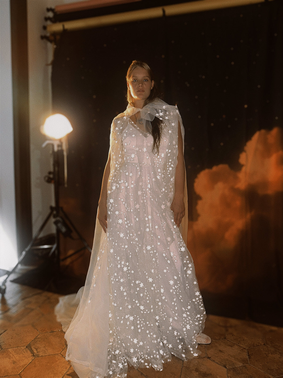Full-length celestial bridal cape with hood and train over a star tulle wedding dress, studio lamp and cloud backdrop, Holly Cape