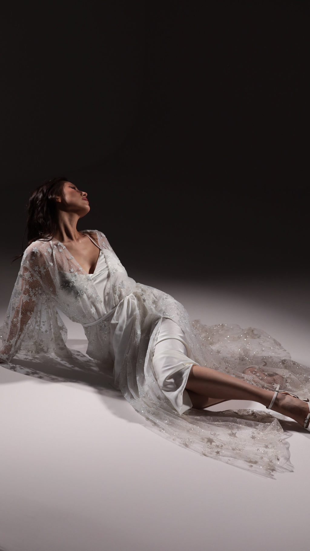 Model reclining on the floor in an ivory slip dress under a star embellished tulle robe spread around her, Infinity Slip Ivory