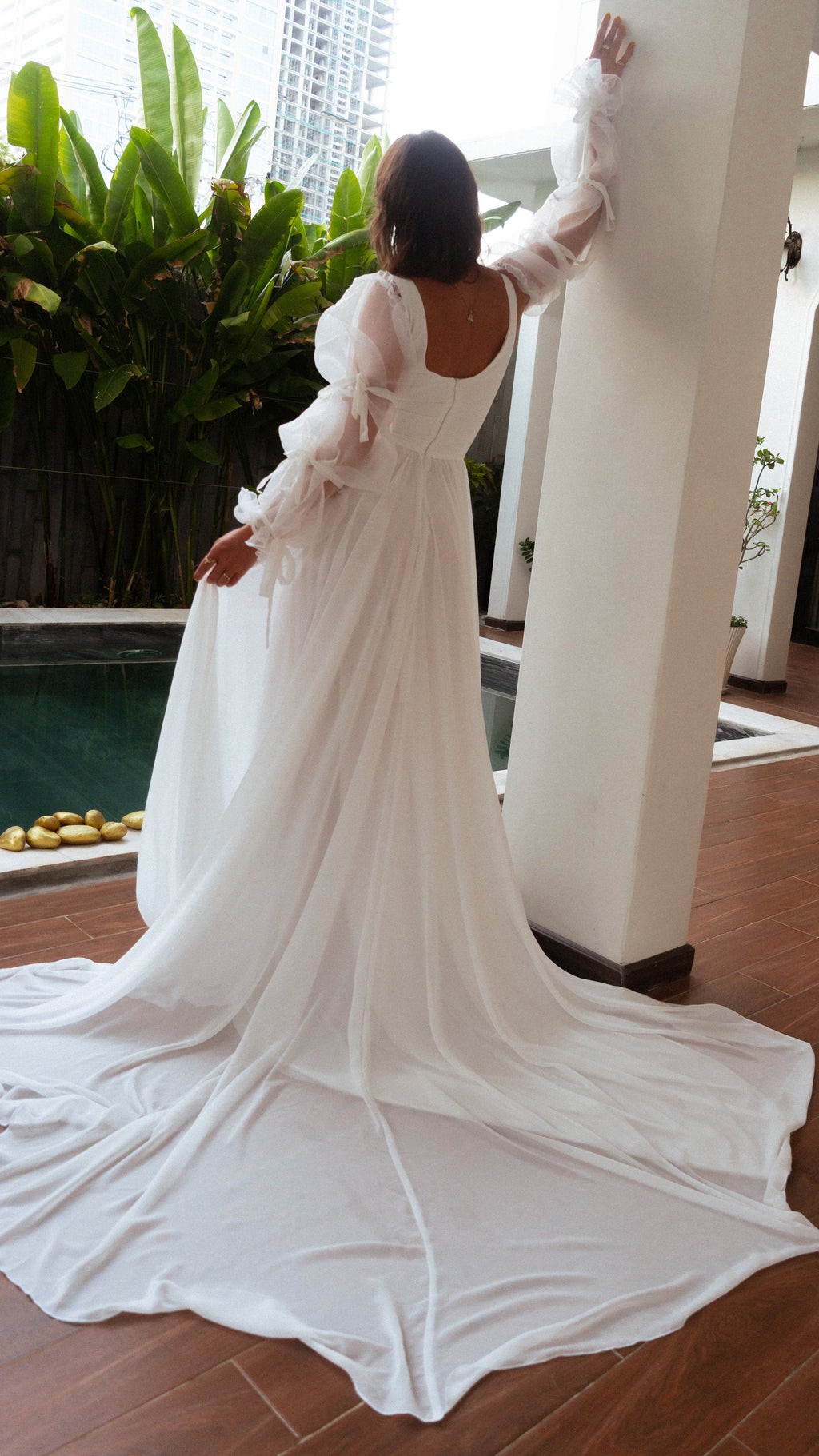 Back view of a bride reaching up a column, square back and long chiffon train of an A-line wedding dress pooling on tiles, LUNA