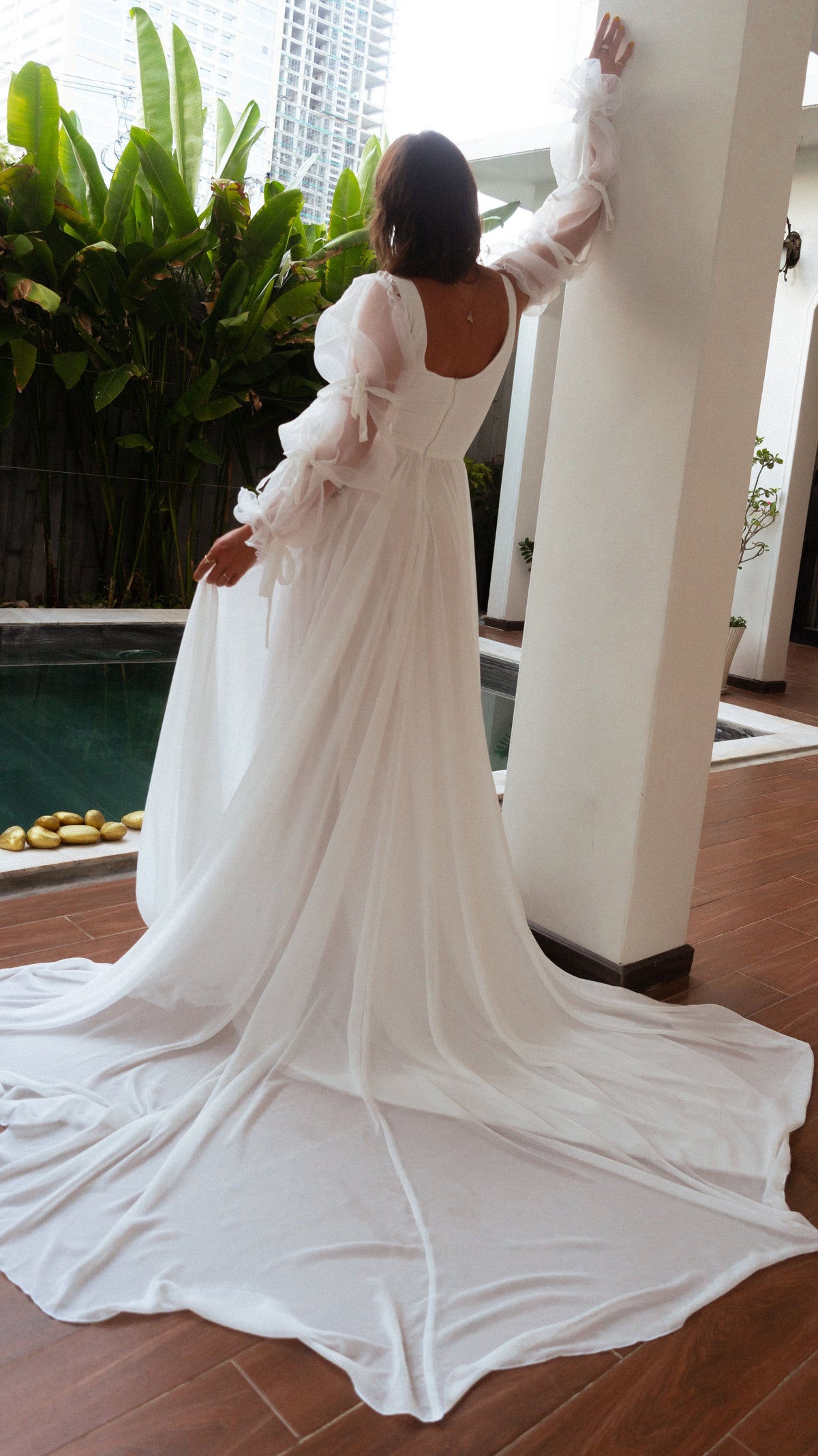 Back view of a bride reaching up a column, square back and long chiffon train of an A-line wedding dress pooling on tiles, LUNA