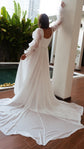 Back view of a bride reaching up a column, square back and long chiffon train of an A-line wedding dress pooling on tiles, LUNA